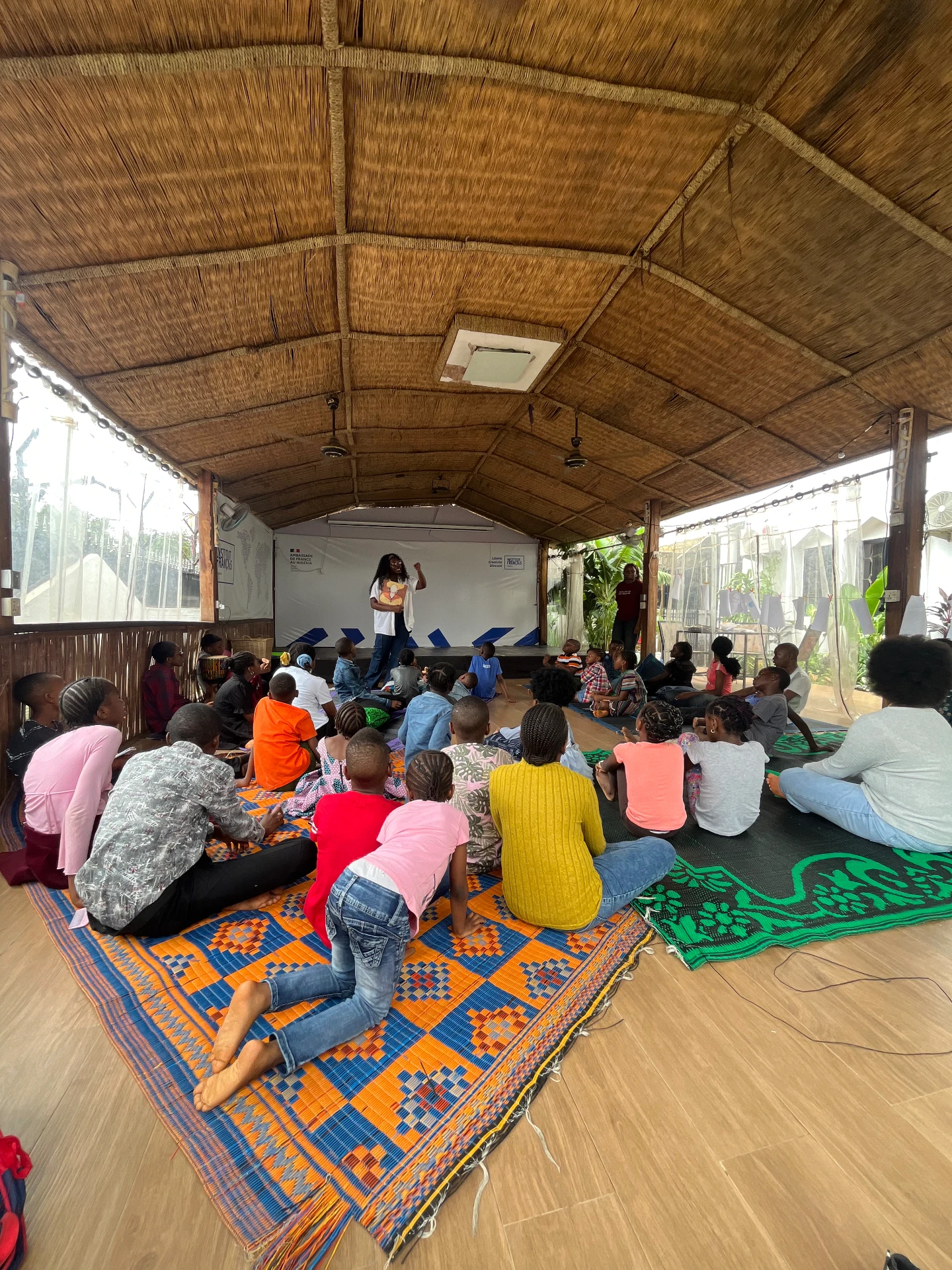 Ibi Kontein leading a children's theatre workshop under a thatched pavilion, with children seated on woven mats