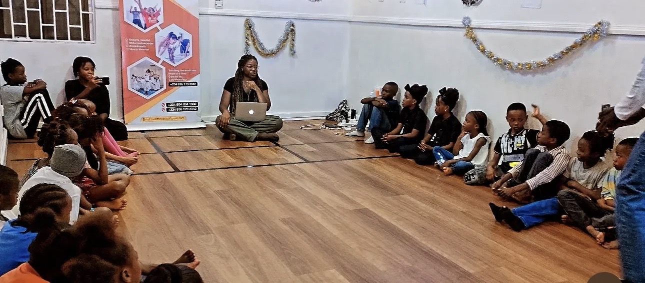 Ibi Kontein leading a children's theatre workshop indoors, with children seated in a semicircle on a wooden floor