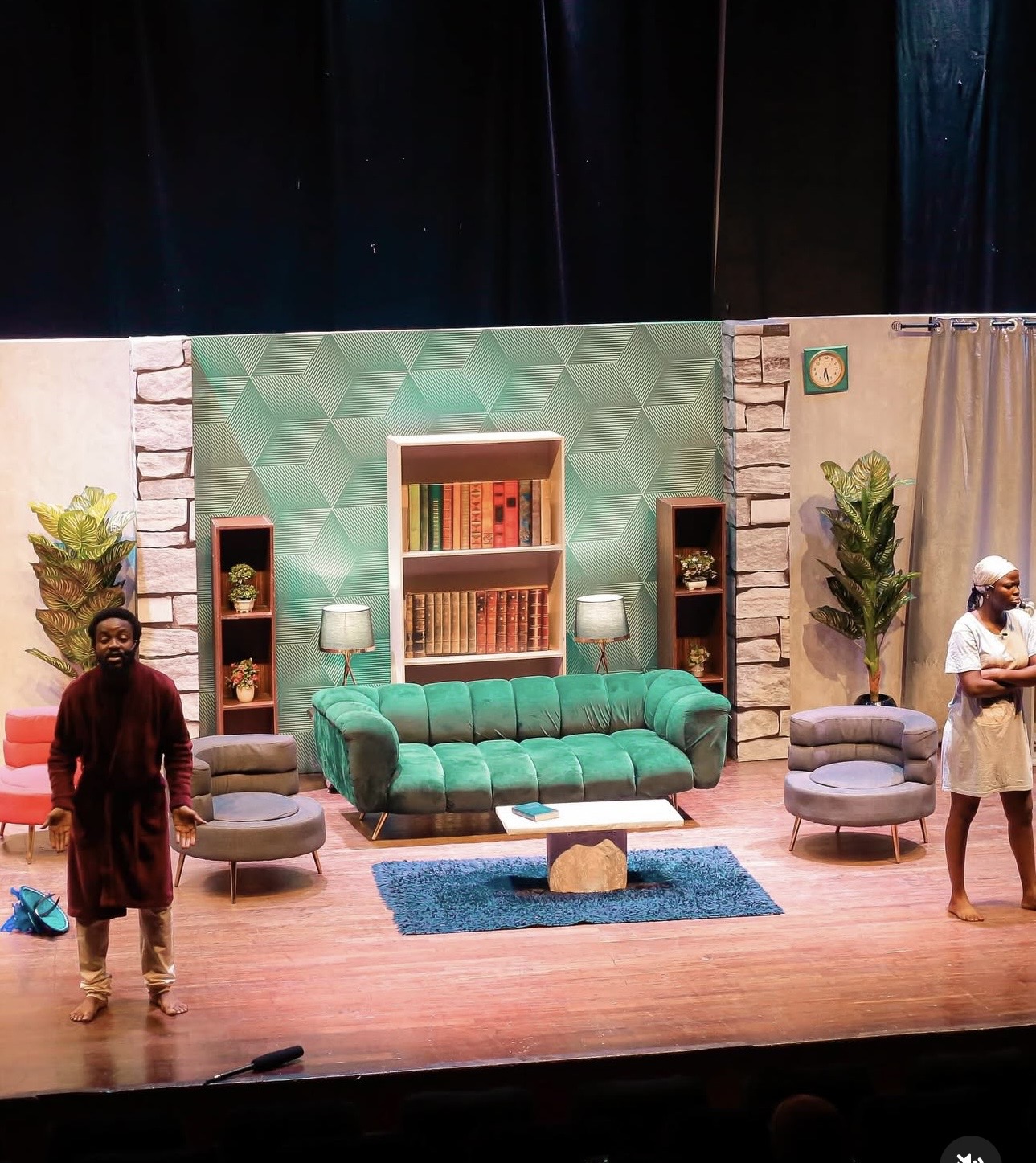 Production photograph from Waterside: two actors on a living-room stage set, one in a burgundy robe with arms outstretched, the other in a white head wrap with arms crossed.